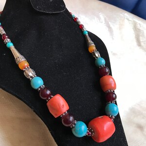 K&ouml;nnte beinhalten: Eine mehrfarbige Perlenkette mit t&uuml;rkisfarbenen, roten, orangefarbenen und silbernen Perlen. Die Kette wird auf einem schwarzen Samtst&auml;nder pr&auml;sentiert.