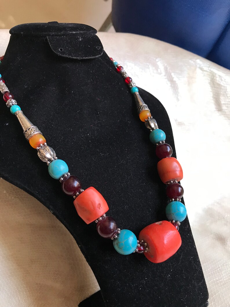 K&ouml;nnte beinhalten: Eine mehrfarbige Perlenkette mit t&uuml;rkisfarbenen, roten, orangefarbenen und silbernen Perlen. Die Kette wird auf einem schwarzen Samtst&auml;nder pr&auml;sentiert.