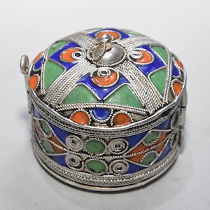 May include: A round, silver-colored trinket box with a hinged lid. The lid and sides are decorated with colorful enamel in blue, green, and orange geometric patterns. The box has a small silver handle on the side and a decorative silver clasp.