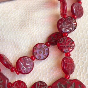 Puede incluir: Un collar de cuentas rojas con cuentas redondas y planas que presentan caligraf&iacute;a &aacute;rabe plateada y dise&ntilde;os de estrellas.