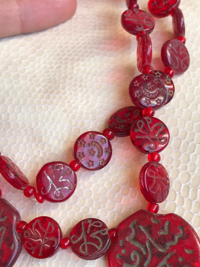 Puede incluir: Un collar de cuentas rojas con cuentas redondas y planas que presentan caligraf&iacute;a &aacute;rabe plateada y dise&ntilde;os de estrellas.