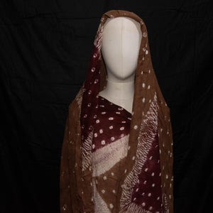 May include: A brown and burgundy scarf with white polka dots and diagonal stripes is draped over a mannequin. The scarf is made of a lightweight fabric and has a draped design. The background is a solid black.