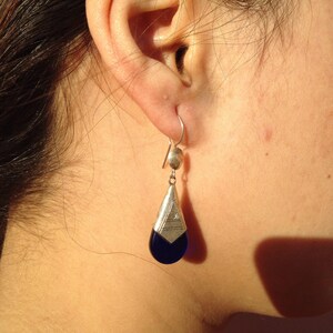 May include: A silver earring with a teardrop-shaped blue stone. The earring has a geometric design at the top and is suspended from a silver hook. The earring is shown on a person's ear.