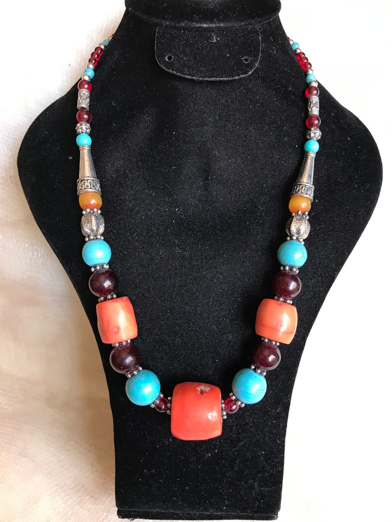 K&ouml;nnte beinhalten: Eine mehrfarbige Perlenkette mit t&uuml;rkisfarbenen, roten und orangefarbenen Perlen. Die Kette hat eine gro&szlig;e, klobige Korallenperle in der Mitte.