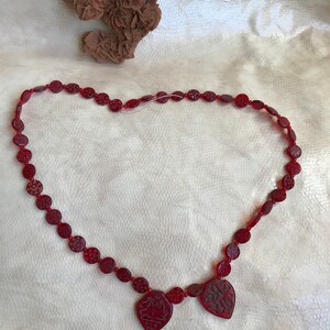 Puede incluir: Un collar de cuentas rojas con tres colgantes en forma de coraz&oacute;n. Los colgantes tienen dise&ntilde;os intrincados tallados en ellos.