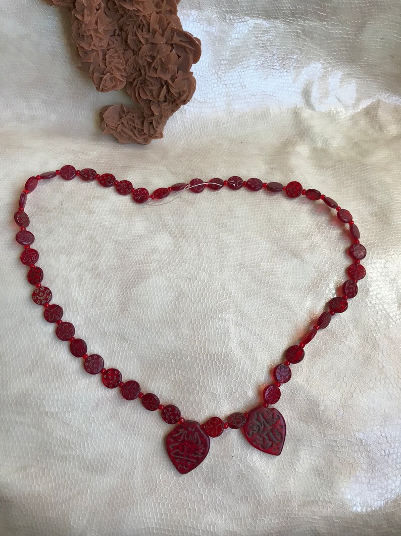 Puede incluir: Un collar de cuentas rojas con tres colgantes en forma de coraz&oacute;n. Los colgantes tienen dise&ntilde;os intrincados tallados en ellos.