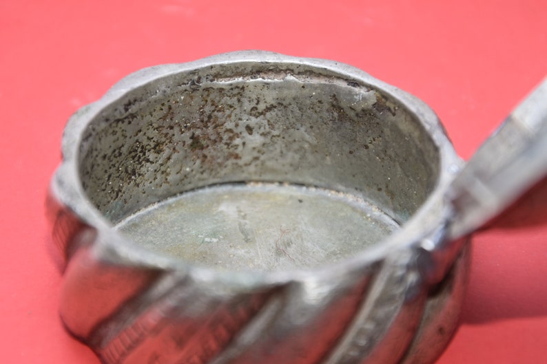 May include: A silver metal container with a twisted design and a lid. The inside of the container is empty and shows signs of wear.