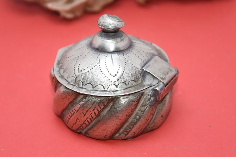May include: A silver trinket box with a hinged lid and intricate engraved designs. The box has a rounded shape and a domed lid with a floral pattern.