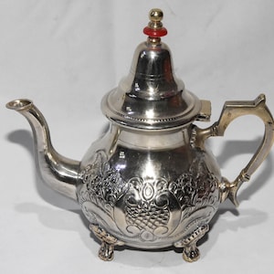 Könnte beinhalten: Silberfarbene Teekanne mit kunstvollen Blumen- und Fruchtmustern. Die Teekanne hat einen gebogenen Ausguss, einen Henkel und einen Deckel mit roter und goldener Verzierung. Die Teekanne steht auf drei kleinen Füßen.