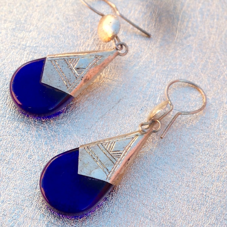 May include: A pair of silver-toned drop earrings with a geometric design. Each earring features a teardrop-shaped, deep blue glass stone, topped with a silver-colored metal piece with etched lines. The earrings have a simple hook closure.