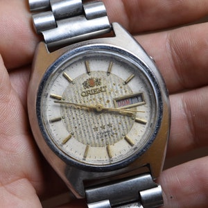 May include: A silver Orient watch with a white face and gold hands. The watch has a day and date window and a silver metal band.