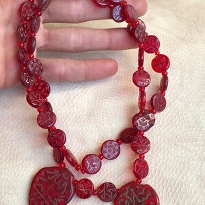 Puede incluir: Un collar de vidrio rojo con dos colgantes en forma de coraz&oacute;n. Las cuentas son redondas y tienen intrincados dise&ntilde;os en oro. Los colgantes tienen caligraf&iacute;a &aacute;rabe en oro.