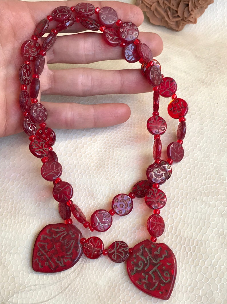 Puede incluir: Un collar de vidrio rojo con dos colgantes en forma de coraz&oacute;n. Las cuentas son redondas y tienen intrincados dise&ntilde;os en oro. Los colgantes tienen caligraf&iacute;a &aacute;rabe en oro.