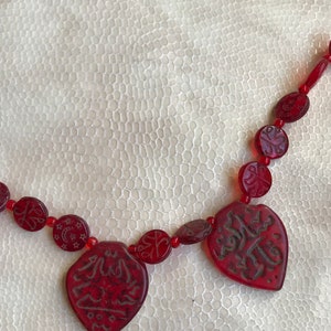Puede incluir: Un collar de cuentas rojas con colgantes tallados en forma de coraz&oacute;n que presentan caligraf&iacute;a &aacute;rabe. El collar est&aacute; sobre una superficie blanca texturizada.