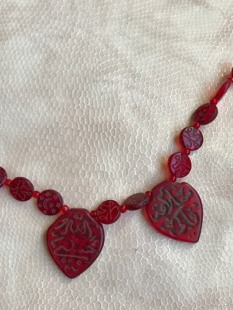 Puede incluir: Un collar de cuentas rojas con colgantes tallados en forma de coraz&oacute;n que presentan caligraf&iacute;a &aacute;rabe. El collar est&aacute; sobre una superficie blanca texturizada.