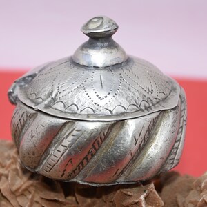May include: A silver trinket box with a decorative lid and a swirling pattern on the body. The lid has a small knob on top.
