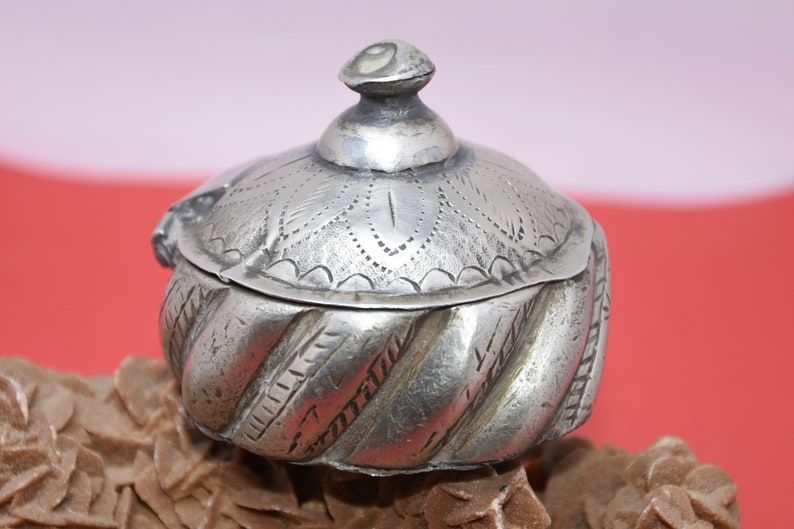 May include: A silver trinket box with a decorative lid and a swirling pattern on the body. The lid has a small knob on top.