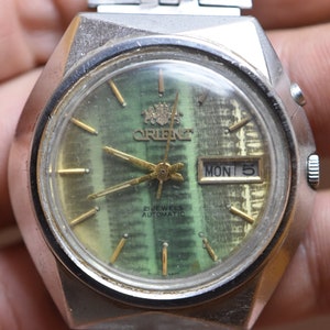 May include: A silver Orient automatic wristwatch with a green face and gold hands. The watch has a date window that displays "MON 5" and the text "21 JEWELS AUTOMATIC" is printed on the face.