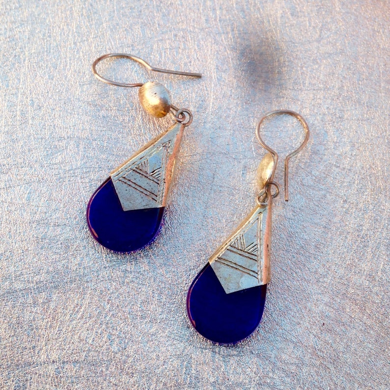 May include: A pair of silver-toned drop earrings with a geometric design. Each earring features a triangular silver top with etched lines and a teardrop-shaped, deep blue glass or stone pendant. The earrings have a simple hook closure.