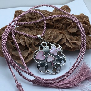 May include: A silver necklace with a pink mother of pearl pendant featuring a heart design. The necklace is on a pink cord with tassels.