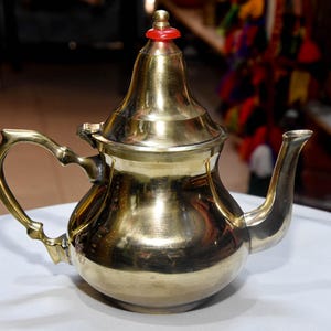 Puede incluir: Una tetera dorada brillante con un asa curva y un pico. La tetera tiene una tapa con un adorno rojo. La tetera está sobre una superficie blanca. Ideal para preparar té.