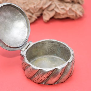 May include: A silver, round, hinged box with a decorative, twisted design. The box is open and the inside is empty.