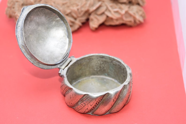 May include: A silver, round, hinged box with a decorative, twisted design. The box is open and the inside is empty.
