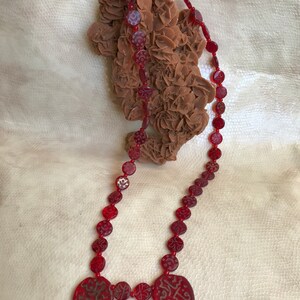 Puede incluir: Un collar de cuentas rojas con dos colgantes en forma de coraz&oacute;n. Las cuentas est&aacute;n talladas con dise&ntilde;os intrincados y los colgantes tienen caligraf&iacute;a &aacute;rabe.