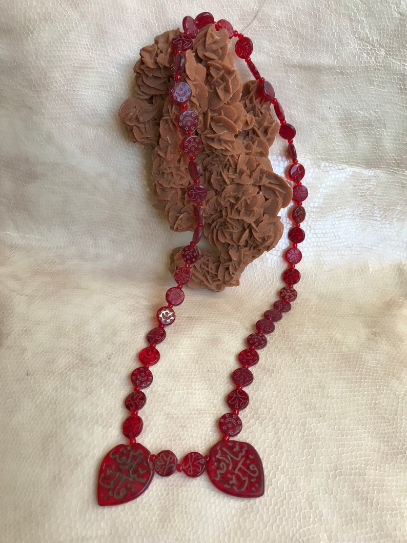 Puede incluir: Un collar de cuentas rojas con dos colgantes en forma de coraz&oacute;n. Las cuentas est&aacute;n talladas con dise&ntilde;os intrincados y los colgantes tienen caligraf&iacute;a &aacute;rabe.