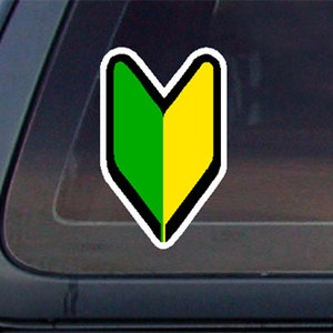 May include: A green and yellow beginner's driver sticker with a white border and black outline. The sticker is shaped like a downward pointing arrow.