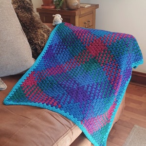 May include: A colourful crocheted blanket with a blue, green, purple and red pattern. The blanket is draped over a brown leather sofa.