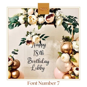 Puede incluir: Un cartel de acrílico transparente con el texto "Happy 18th Birthday Libby" en letra cursiva negra. El cartel está decorado con un pequeño ramo de flores blancas y rosas. El cartel está rodeado de un arco de globos dorados, blancos y rosas.