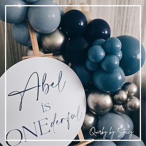 Puede incluir: Un cartel redondo blanco con el texto "Abel is ONEderful" en letra cursiva negra. El cartel está apoyado en un caballete de madera. El cartel está rodeado de una guirnalda de globos azules, plateados y crema.