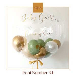 Może przedstawiać: Przezroczysty balon z napisem "Baby Gartshore Coming Soon" w kolorze złotym. W środku mniejsze balony w odcieniach złota, zieleni i bieli. Złota wstążka u dołu. Tekst "Font Number 34" na dole.