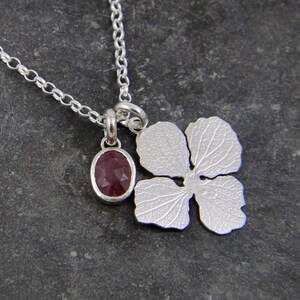 Puede incluir: Un collar de plata con una cadena delicada y un colgante con una pequeña piedra preciosa de color rojo oscuro de forma ovalada y un dije de flor de plata.