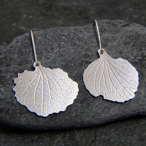 May include: A pair of silver earrings shaped like leaves with detailed veins. The earrings are hanging from a silver hook.