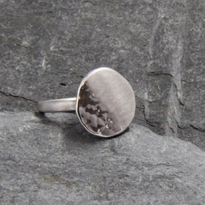 May include: A silver ring with a hammered, circular face. The ring is sitting on a gray rock.