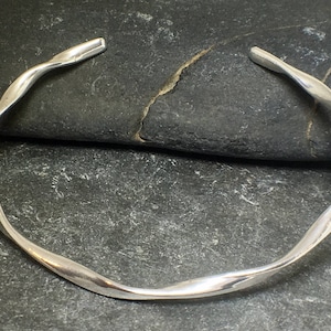 May include: A silver bangle bracelet with a twisted, wavy design. The bracelet is open and has a smooth, polished finish.