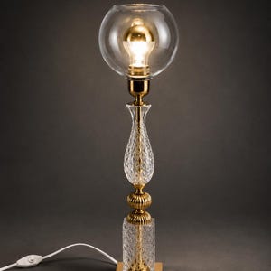 Peut inclure: Une lampe de table décorative avec un abat-jour en forme de globe en verre transparent et une ampoule dorée. La lampe présente une base et des accents dorés, avec une colonne en verre clair et texturé. La lampe est connectée à un cordon d'alimentation blanc.
