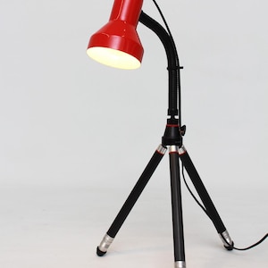 Könnte beinhalten: Eine rote Schreibtischlampe mit einem schwarzen, flexiblen Hals und einem Stativfuß. Der Lampenschirm ist leuchtend rot, und der Fuß hat silberne Akzente. Die Lampe ist für fokussiertes Licht konzipiert und hat eine moderne Ästhetik.