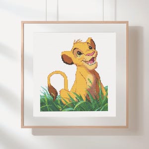 Puede incluir: Arte de punto de cruz de Simba de El Rey León. La obra presenta un cachorro de león amarillo sonriente sentado en hierba verde. La pieza enmarcada tiene un marco de madera clara y está colgada en una pared blanca.