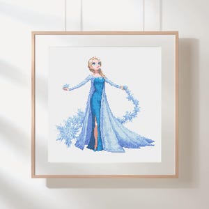 Peut inclure: Une œuvre d'art au point de croix encadrée, représentant un personnage en robe bleue longue fendue. Le personnage a les cheveux blonds et est entouré de détails sur le thème des flocons de neige et de la glace. Le cadre est en bois clair.