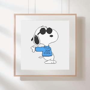 May include: A framed cross-stitch featuring Snoopy wearing sunglasses and a blue sweater with the text "JOE COOL". The artwork is in a pixelated style, with a white background and a wooden frame. The image is hanging on a white wall.