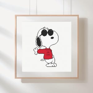 May include: Framed cross-stitch art featuring Snoopy wearing sunglasses and a red sweater with "JOE COOL" text. The artwork is displayed in a light-colored wooden frame, perfect for a child's room or a Peanuts fan.