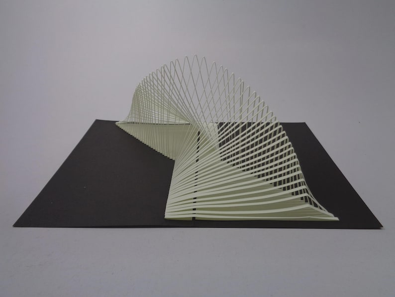 May include: A white paper sculpture with a curved, wave-like design. The sculpture is composed of numerous parallel lines, creating a three-dimensional effect. It sits on a black rectangular base against a gray background.