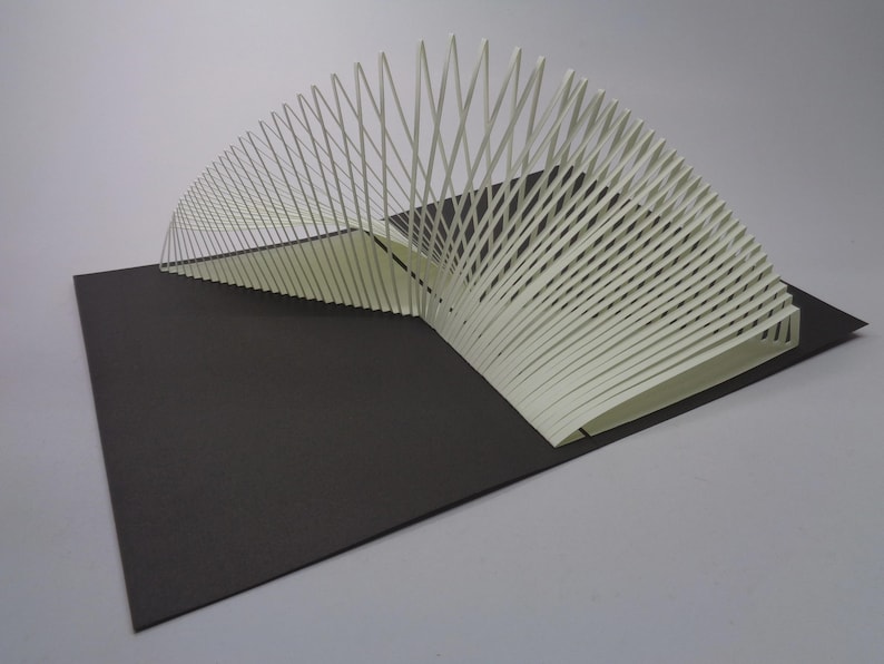 May include: A white paper sculpture with a fan-like design, composed of numerous vertical lines, sits on a dark gray rectangular base. The sculpture's lines curve gracefully, creating a sense of depth and dimension. The artwork is set against a neutral background.