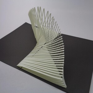 May include: A white paper sculpture with a spiral design, composed of numerous parallel strips, stands on a dark gray rectangular base. The sculpture curves upwards, creating a dynamic visual effect. The background is a plain white surface.