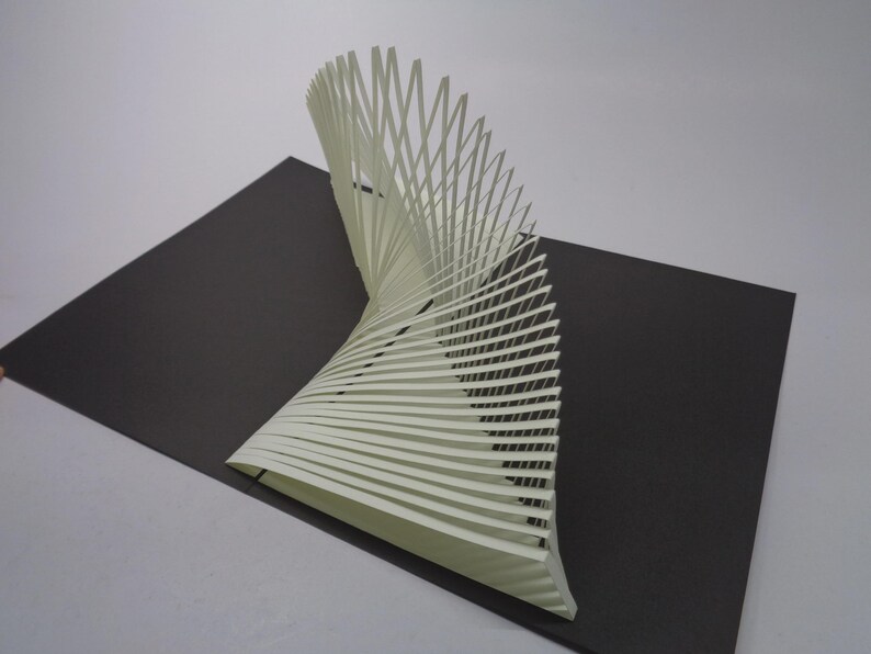 May include: A white paper sculpture with a spiral design, composed of numerous parallel strips, stands on a dark gray rectangular base. The sculpture curves upwards, creating a dynamic visual effect. The background is a plain white surface.