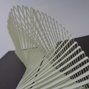 May include: A cream-colored, abstract sculpture with a spiral design. The artwork is composed of numerous thin, curved strips arranged in a repeating pattern, creating a sense of depth and movement. The background is a mix of gray and black.