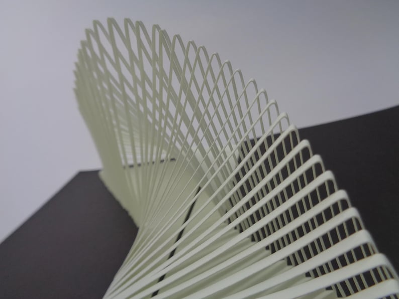 May include: A cream-colored, abstract sculpture with a spiral design. The artwork is composed of numerous thin, curved strips arranged in a repeating pattern, creating a sense of depth and movement. The background is a mix of gray and black.
